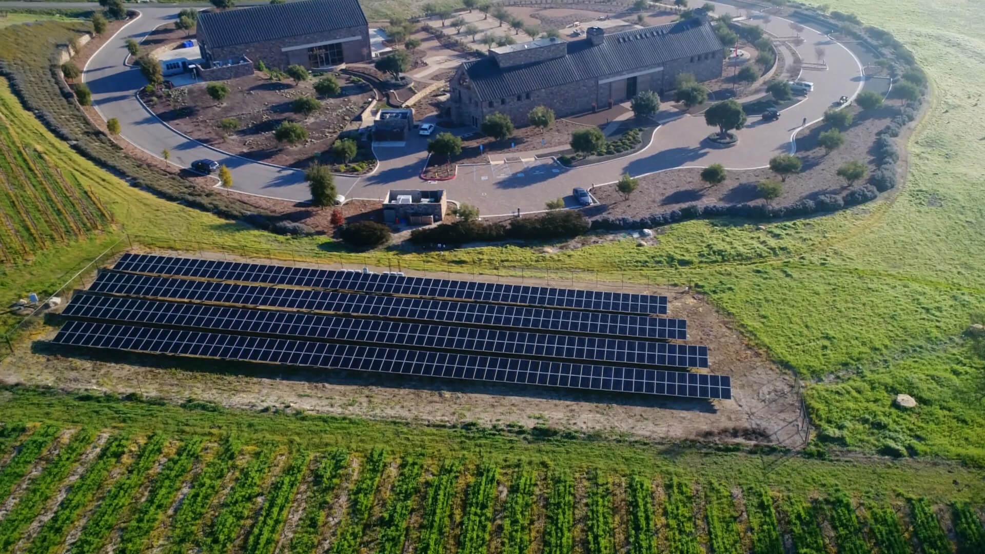 A drone shot showing a green landscape, with solar panels in a field and a stone tasting room.