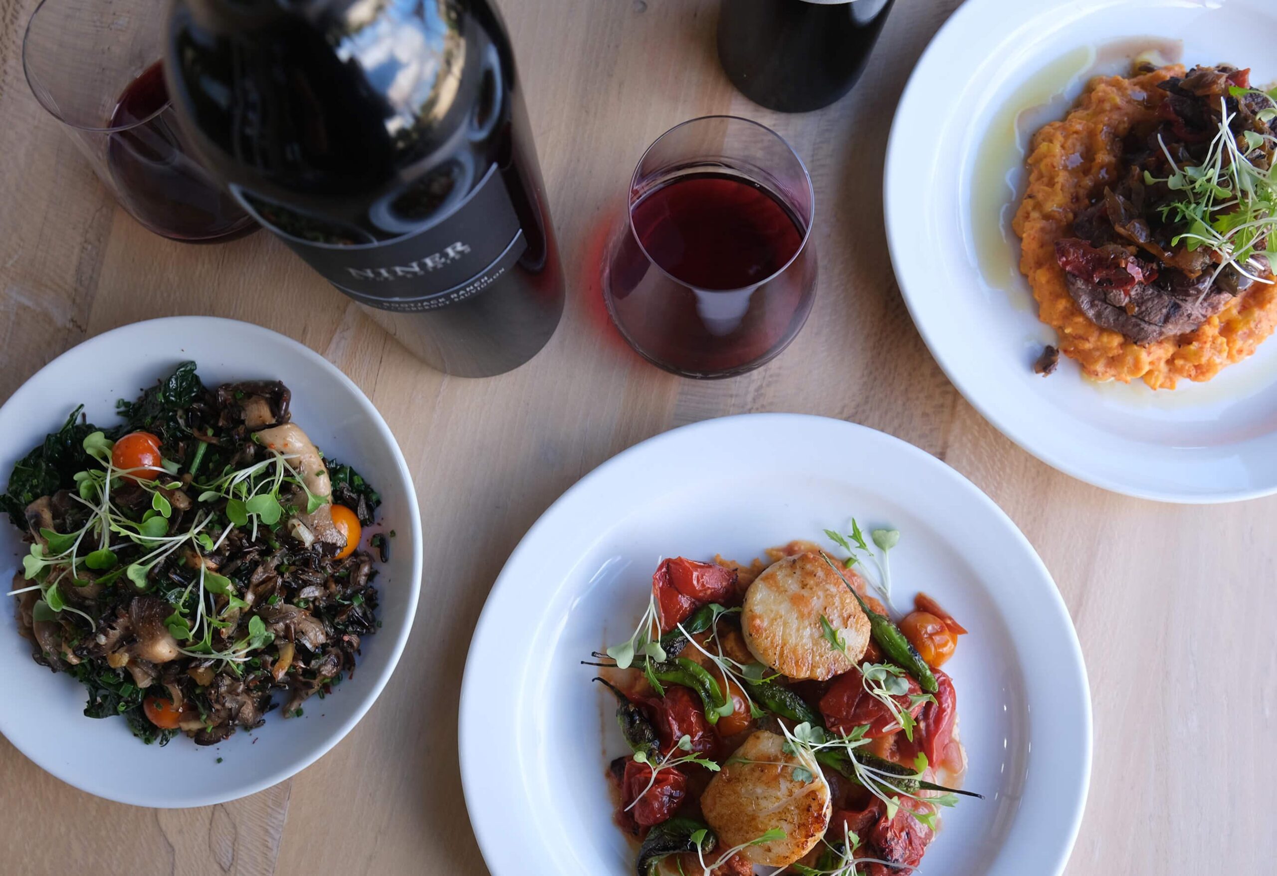 An array of colorful dishes and a bottle and glass of wine on a table, including scallops, steak and wild rice.